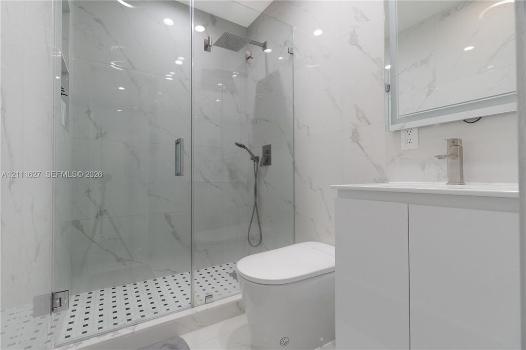 Walk-in glass-enclosed shower with a rainfall showerhead, large-format tile surround, mosaic tile flooring, and integrated wall niche, complemented by a white vanity with a metallic faucet and integrated lighting