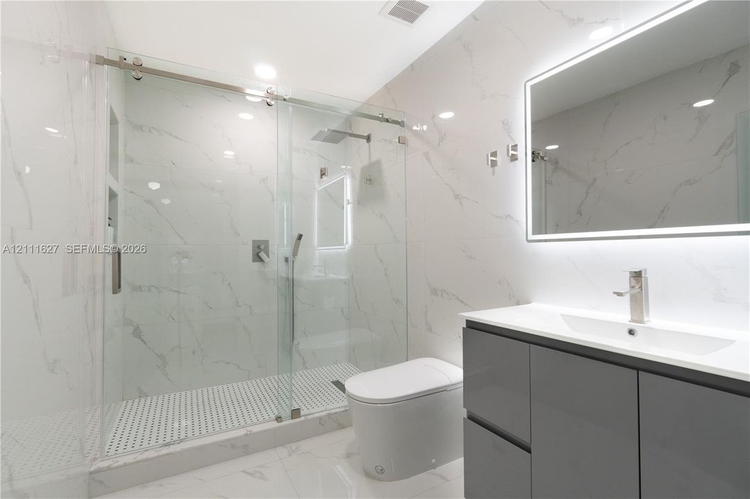 Walk-in glass-enclosed shower featuring stone-finish wall tile, rainfall showerhead, and built-in recessed shelving, complemented by a modern floating vanity with integrated lighting and a streamlined toilet