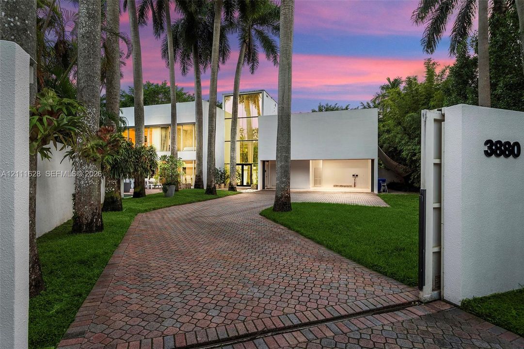 Modern architectural exterior featuring a structured smooth-stucco facade, expansive floor-to-ceiling glass paneling, a multi-car garage, and a gated entry with a patterned stone paver driveway