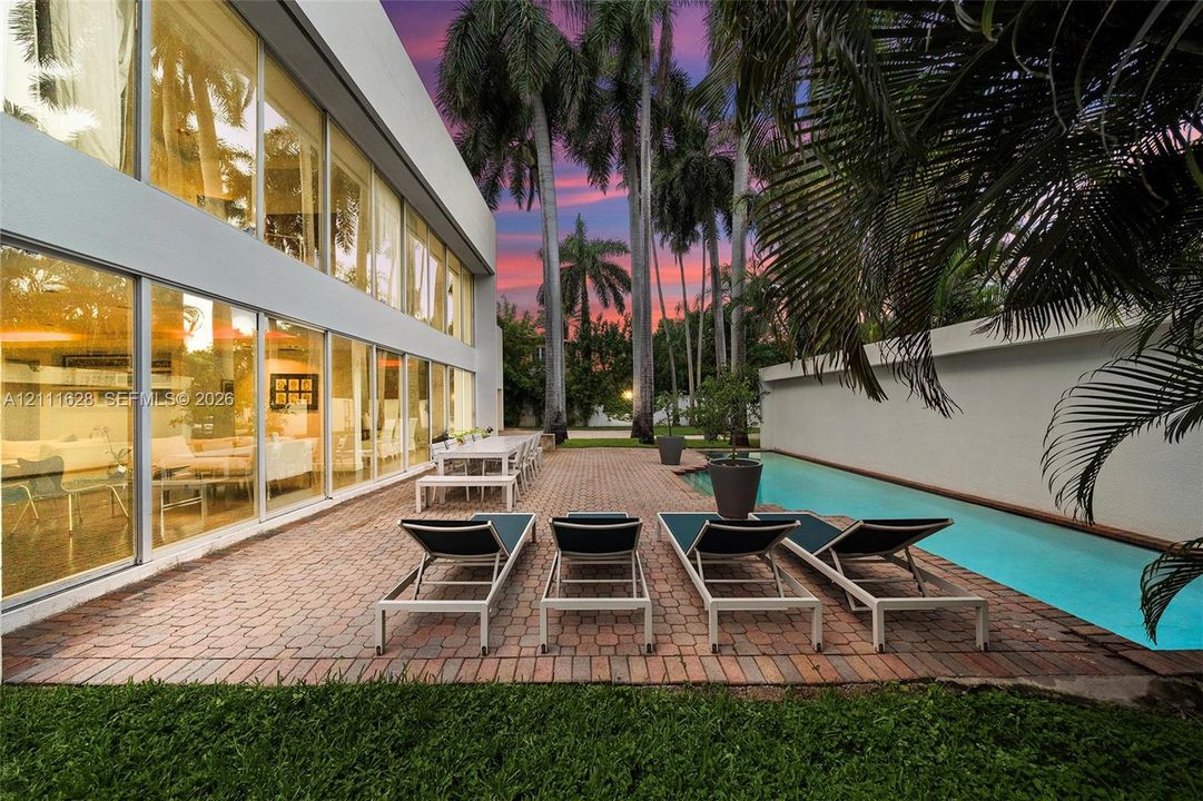 Outdoor patio featuring brick pavers, floor-to-ceiling glass paneling, a lap pool, and mature palm trees