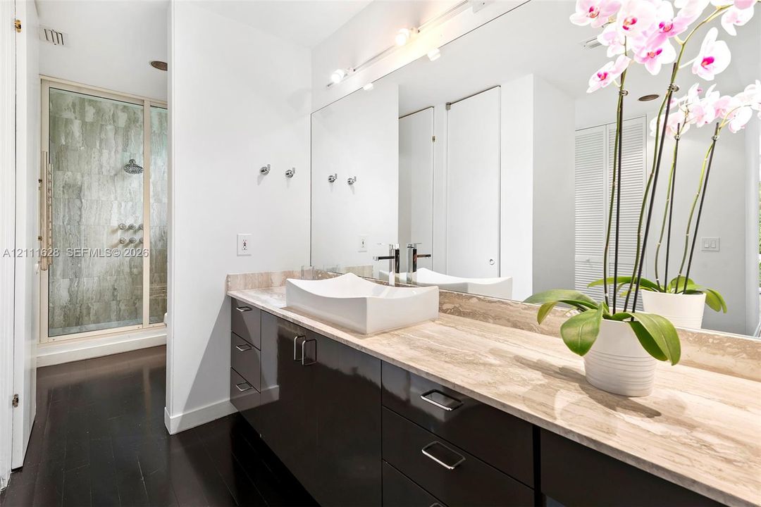 Bathroom featuring a natural stone countertop, vessel sink with chrome faucet, dark-finish cabinetry, floor-to-ceiling mirror, and a walk-in shower with stone tile surround