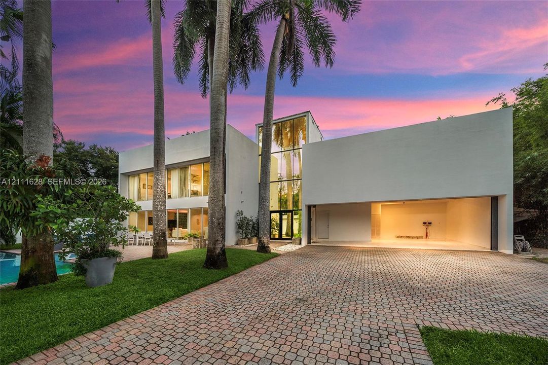 Contemporary exterior featuring multi-level floor-to-ceiling glass windows, a two-car open-bay garage, paver driveway, and mature palm trees