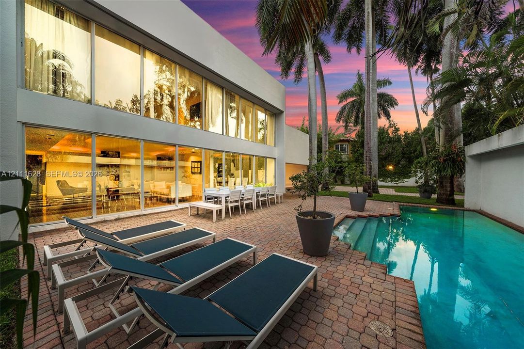 Modern exterior facade featuring floor-to-ceiling glass paneling, private swimming pool, paver patio, and mature palm trees