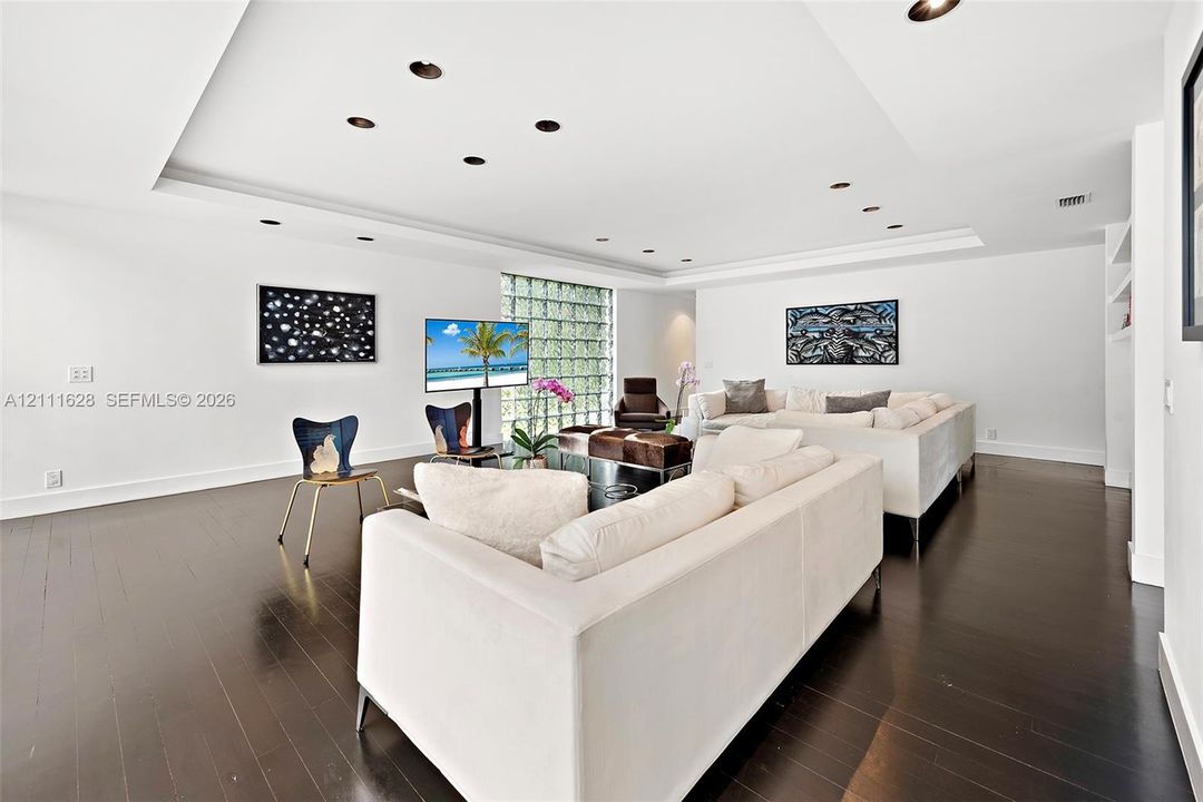 Open-concept living space featuring wood-finish flooring, a recessed tray ceiling, integrated recessed lighting, glass block accent wall, and built-in shelving