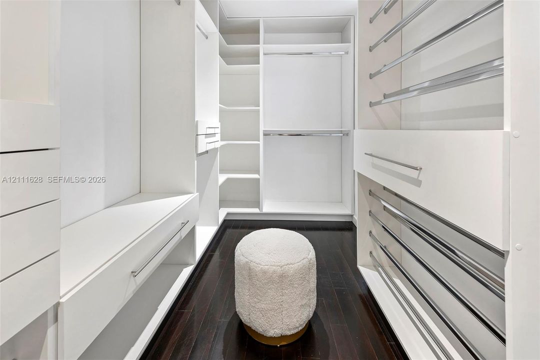 Custom walk-in closet featuring white built-in shelving, integrated drawers, polished metal hanging rods, and dark wood-finish flooring