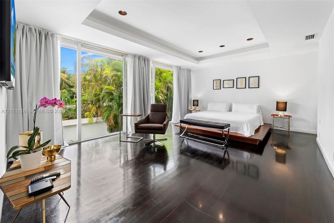 Floor-to-ceiling glass sliding doors, polished dark wood-finish flooring, tray ceiling with recessed lighting, and expansive exterior foliage