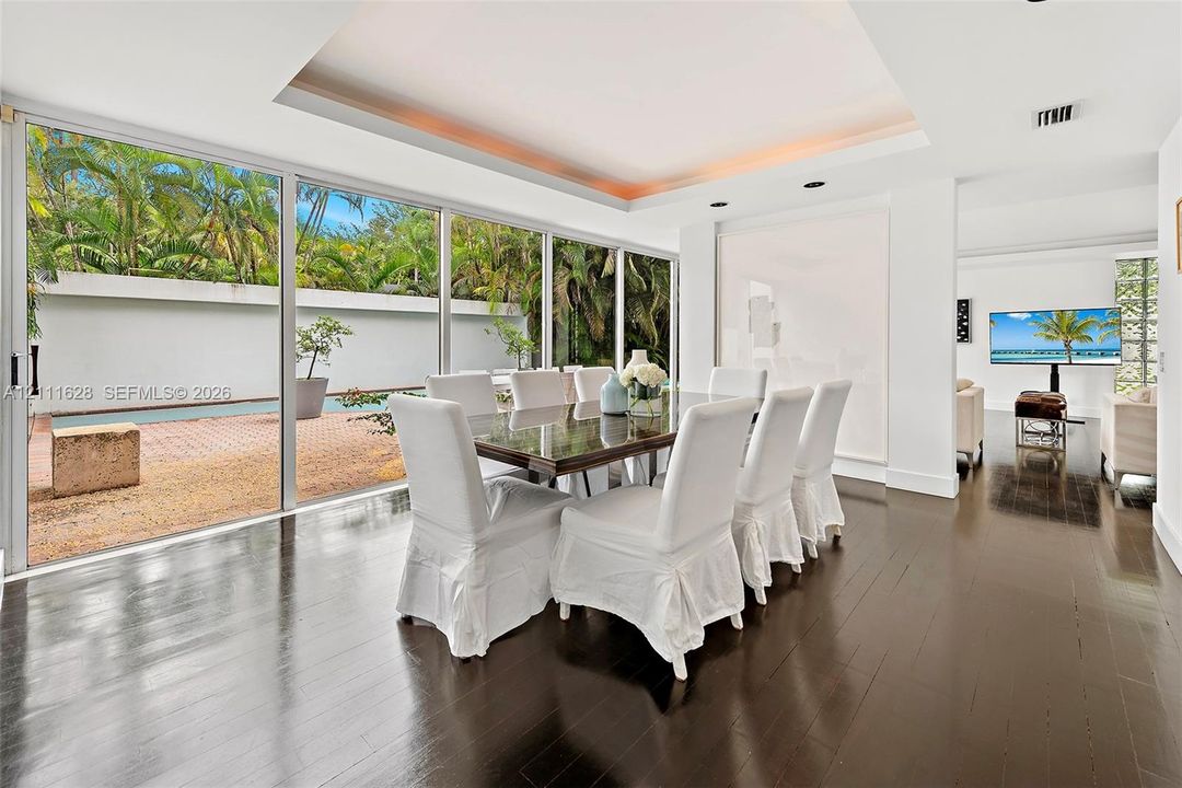 Floor-to-ceiling sliding glass walls, dark wood-finish flooring, recessed tray ceiling with integrated lighting, and a paved outdoor patio area