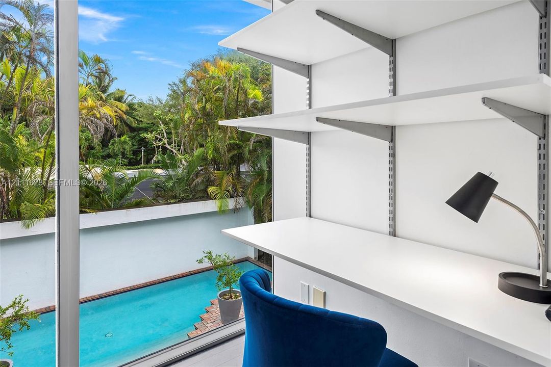Built-in white shelving system with integrated desk surface, expansive floor-to-ceiling glass panel, and exterior swimming pool
