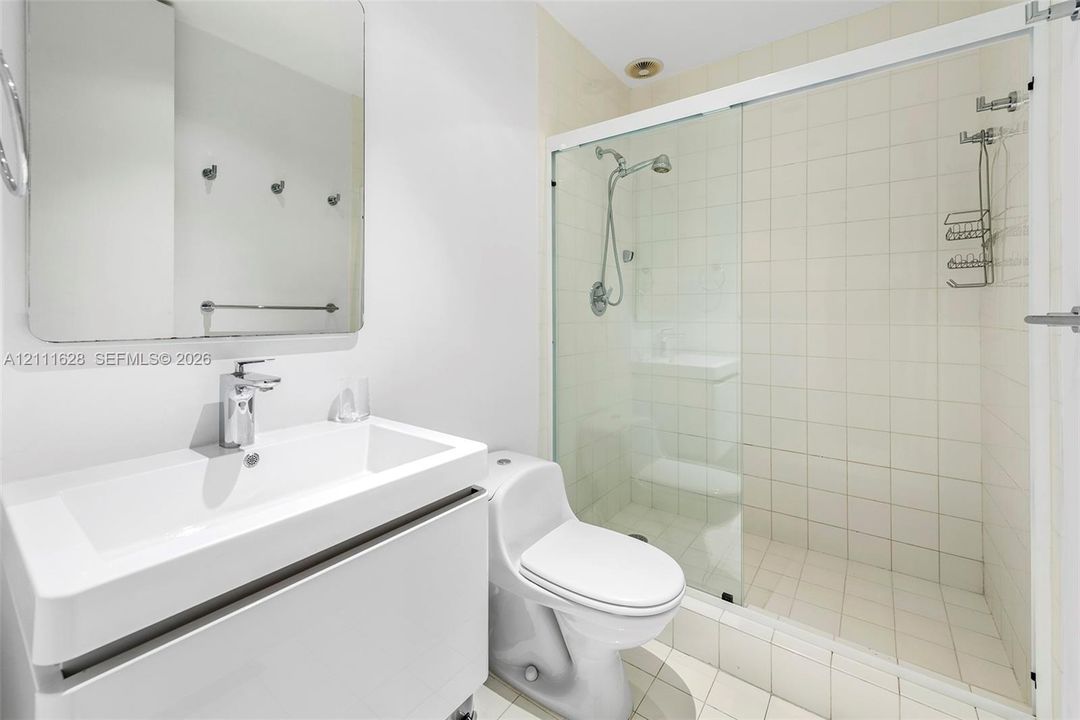 White vanity with integrated rectangular sink, wall-mounted mirror, glass-enclosed shower featuring tiled walls and flooring, and a low-profile toilet