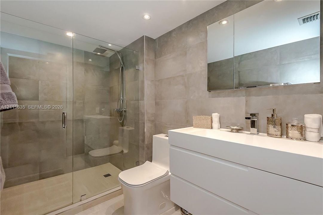 Bathroom featuring large-format neutral wall tile, walk-in glass shower enclosure with rainfall showerhead, white wall-mounted vanity, undermount sink with chrome faucet, and rectangular medicine cabinet