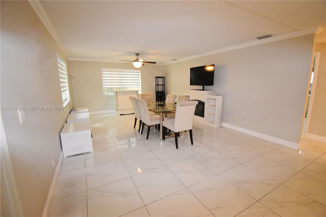 Large-format tile flooring, white built-in fireplace mantle, crown molding, ceiling fan, and window blinds