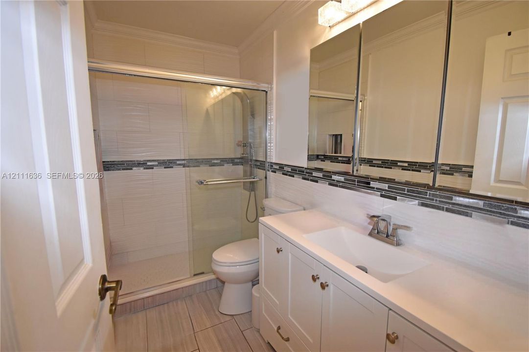 Bathroom featuring a vanity with a white countertop and integrated sink, a glass-enclosed shower with tiled surround, stone accent tile detailing, mirrored medicine cabinetry, and light-toned tile flooring