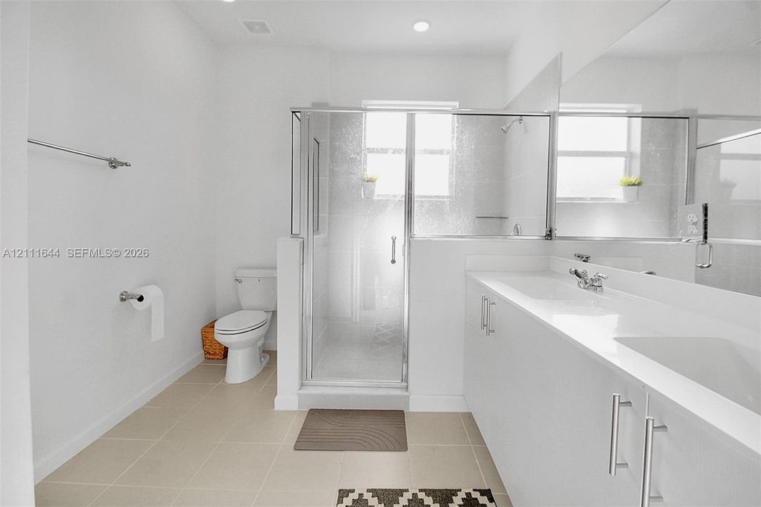 Dual-sink vanity with white cabinetry and solid surface countertop, glass-enclosed shower, polished chrome fixtures, and neutral floor tiling