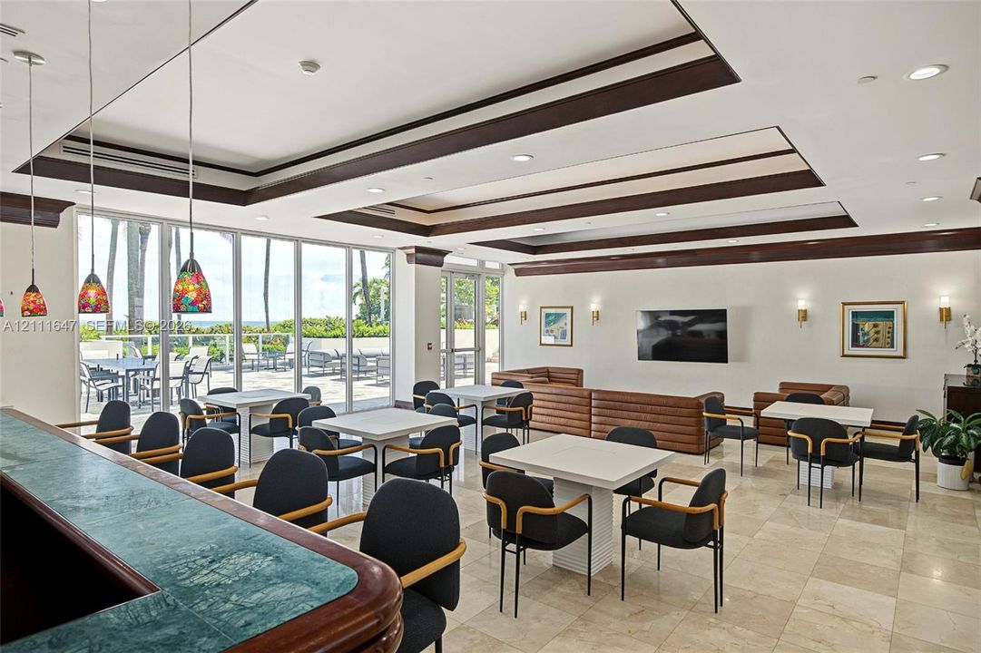 Spacious common area featuring recessed tray ceilings with dark wood trim, tile flooring, built-in stone-top bar, and expansive floor-to-ceiling glass wall panels