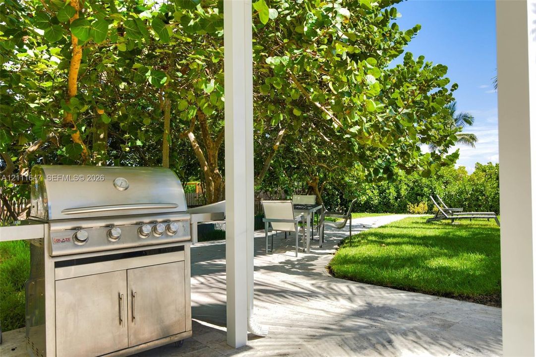 Covered patio featuring a built-in stainless steel grill, stone paver flooring, and mature landscaping surrounding a grass lawn