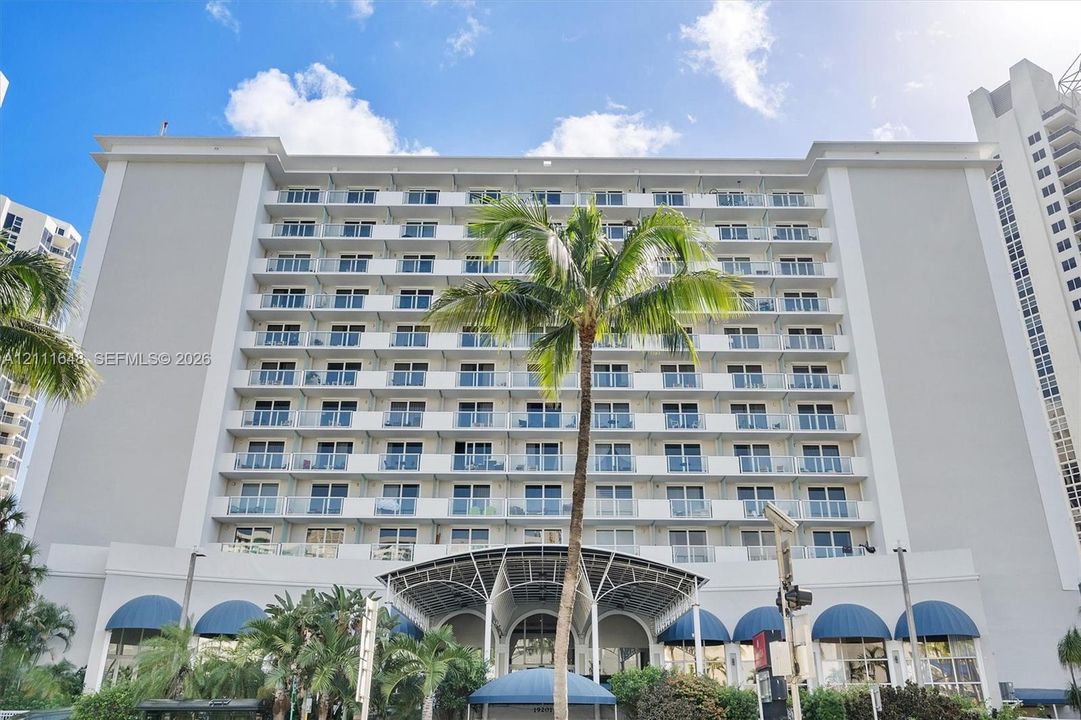 Mid-rise building exterior featuring multiple levels of private balconies with glass railings, a prominent arched entrance canopy, and blue architectural awnings