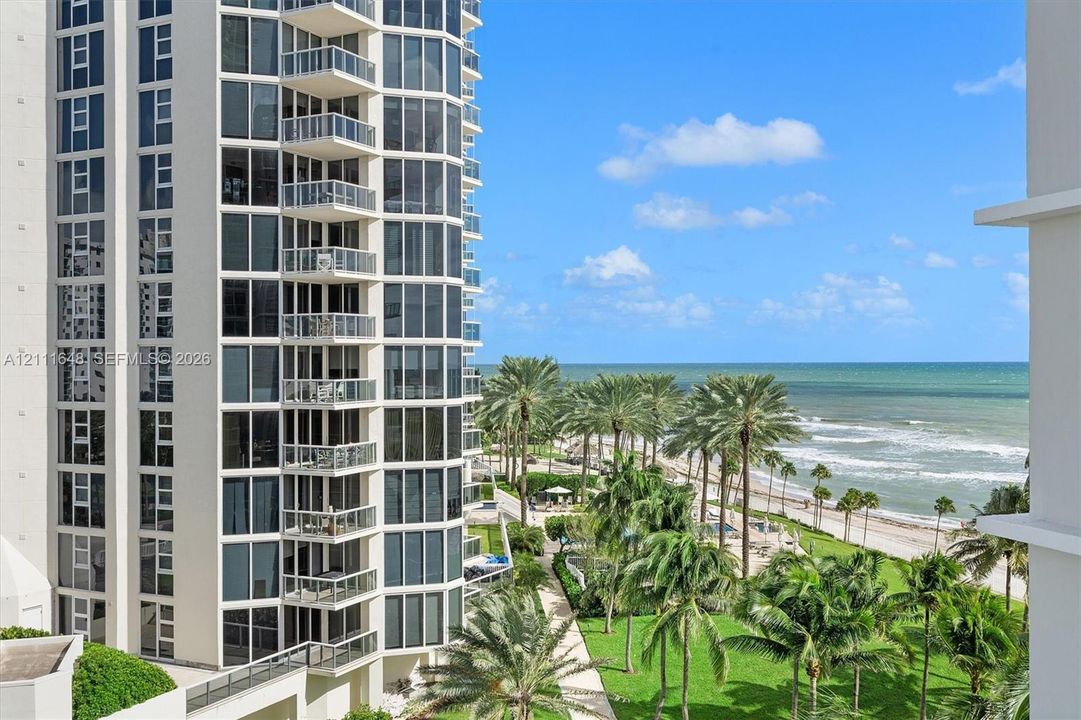 High-rise tower featuring floor-to-ceiling glass windows and stacked private balconies, set against an oceanfront backdrop with palm-lined landscaping
