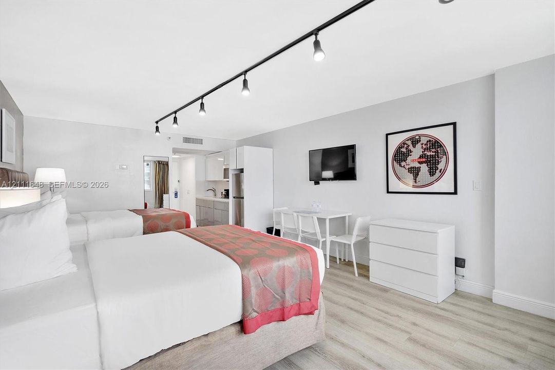 Light-toned wood-finish flooring, linear track lighting, and a kitchenette featuring white cabinetry and grey countertops