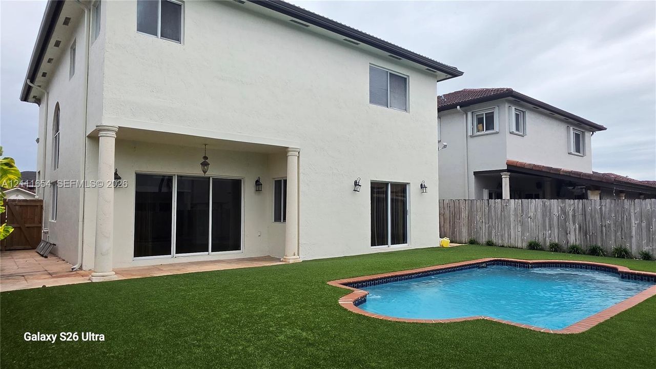 Two-story stucco exterior featuring a covered patio with architectural columns, synthetic turf lawn, in-ground swimming pool with brick coping, and wood privacy fencing