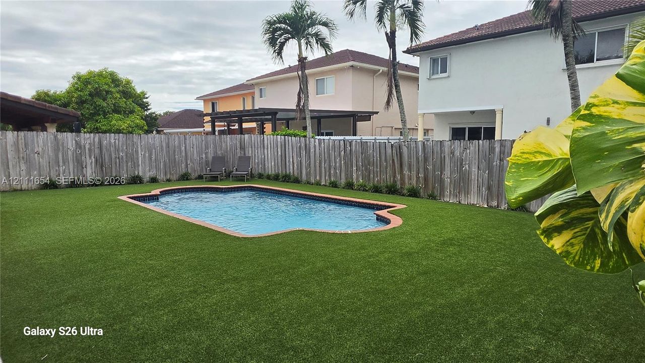 Backyard area featuring an in-ground swimming pool with brick-style coping, synthetic turf lawn, and a perimeter wood privacy fence