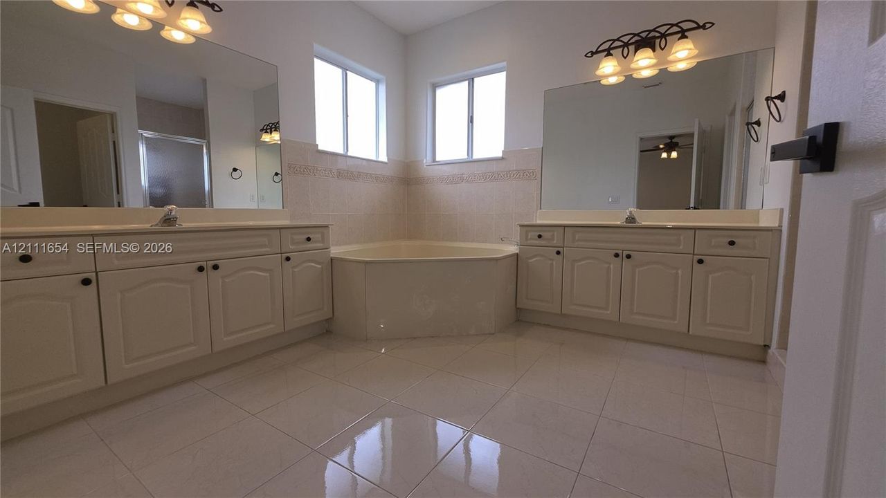 Dual vanities featuring raised-panel cabinetry, corner soaking tub with tiled surround, large wall-mounted mirrors, and polished ceramic tile flooring