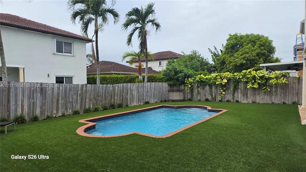 Private backyard featuring a swimming pool with a red-brick border, synthetic turf, wood-plank fencing, and mature palm trees