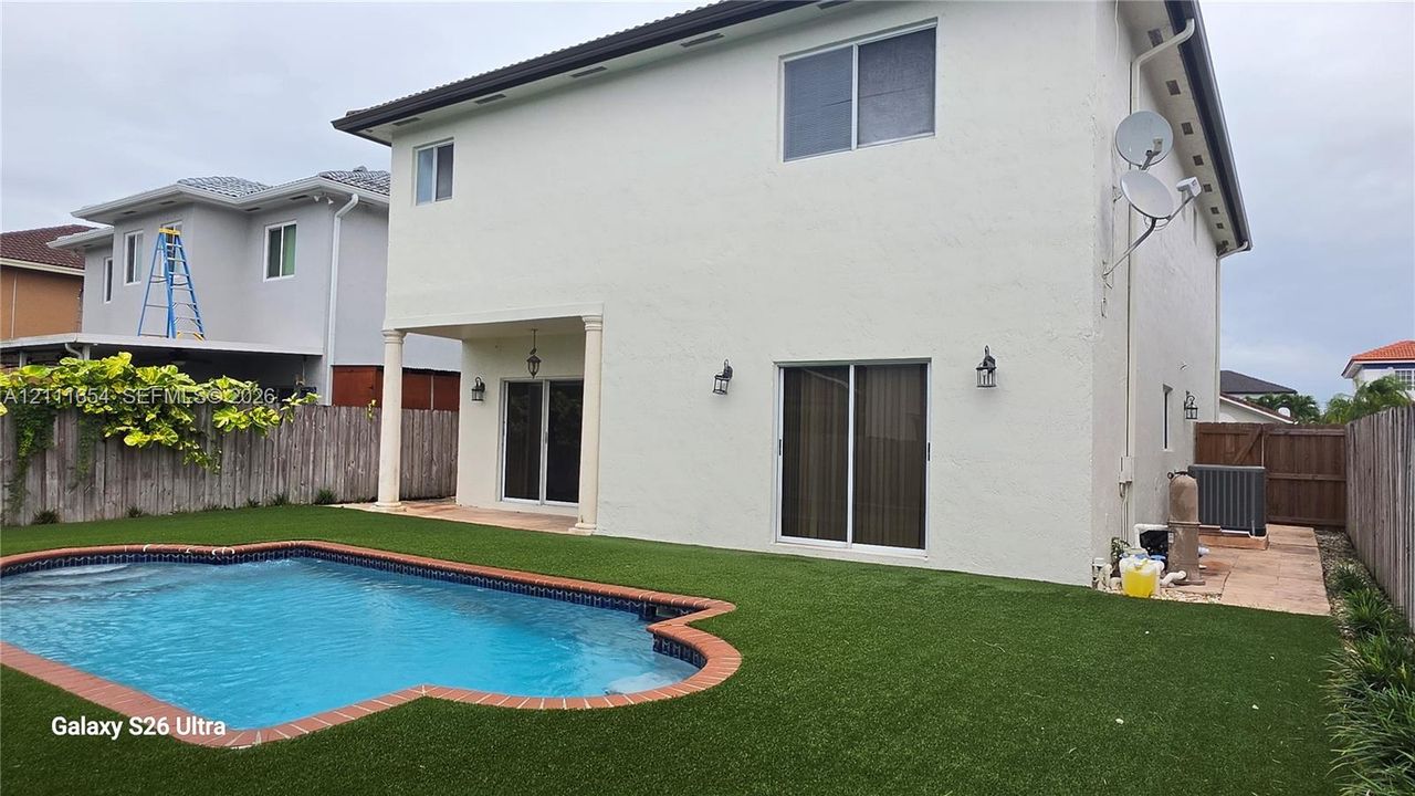Two-story exterior featuring a stucco facade, covered patio with structural columns, synthetic turf landscaping, and an in-ground swimming pool with brick-style coping