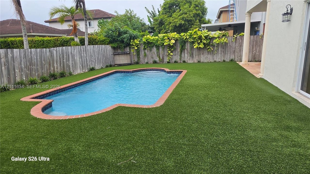 Private backyard featuring a swimming pool with brick coping, synthetic turf lawn, and perimeter wood privacy fencing