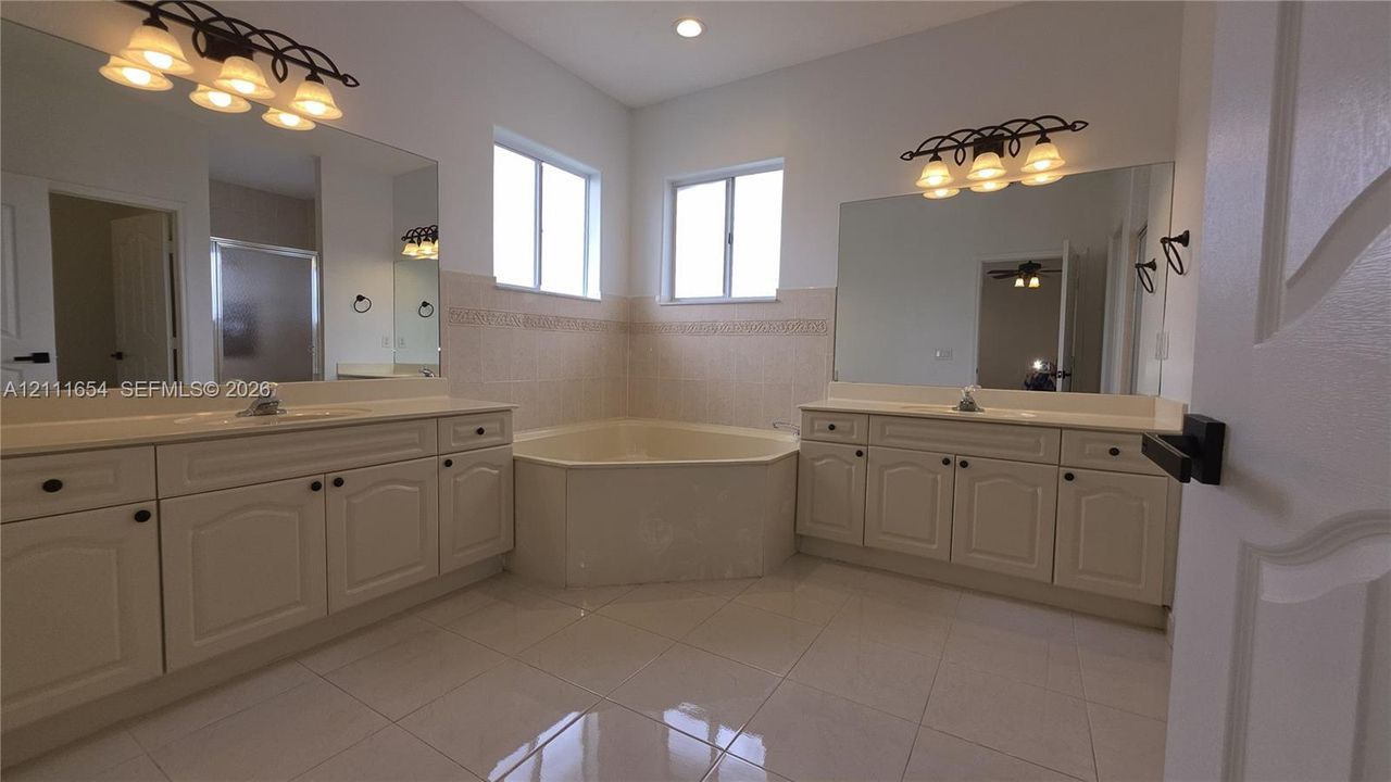 Dual vanity cabinetry with solid surface countertops, a built-in corner soaking tub with tile surround, large wall-mounted mirrors, and tiled flooring