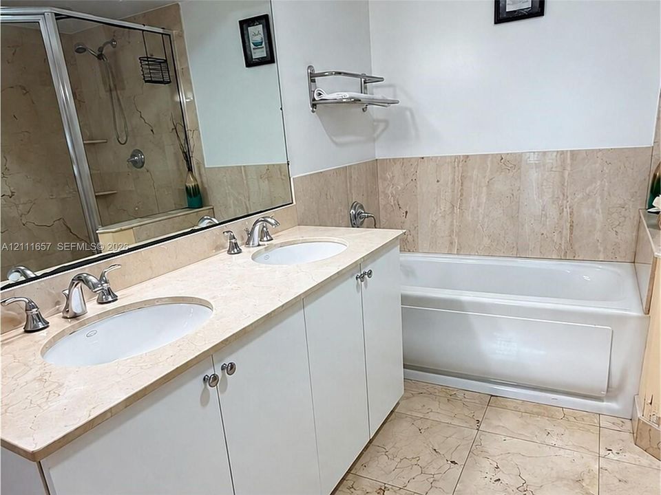 Dual-sink vanity with stone countertop, drop-in soaking tub with stone surround, tile flooring, wall-to-wall mirror, and glass-enclosed shower