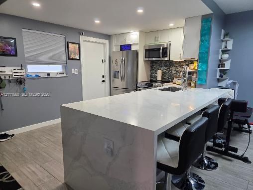 Kitchen featuring a waterfall island with stone-finish countertop, stainless steel appliances, mosaic tile backsplash, white cabinetry, and ceramic-finish flooring