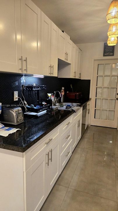 Kitchen featuring white shaker-style cabinetry, dark stone countertops, a textured black backsplash, large-format tile flooring, and a glass-paneled door