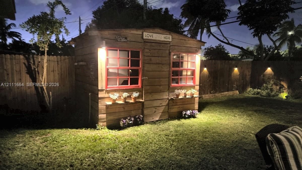 Detached wood-sided structure featuring two windows with red frames, built-in exterior shelving, wall-mounted lighting, and a fenced backyard