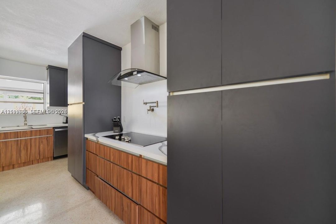 Kitchen featuring custom flat-panel cabinetry with wood-finish lower cabinets and matte charcoal upper cabinetry, stainless steel range hood, integrated cooktop, and wall-mounted pot filler faucet