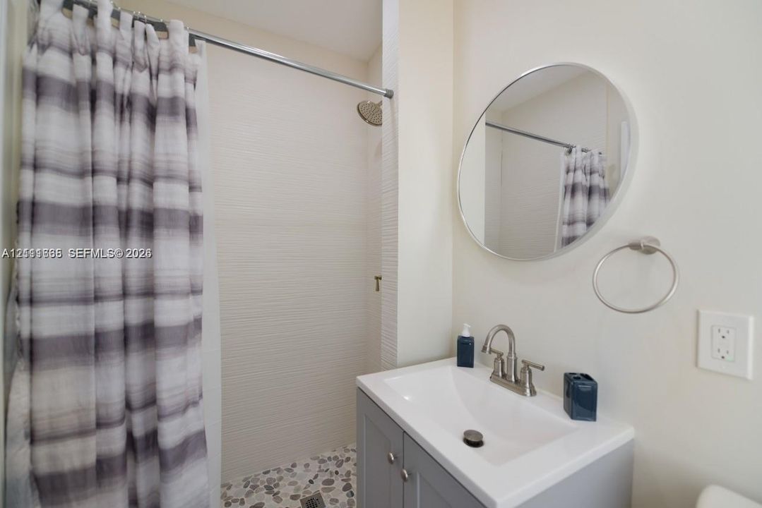 Bathroom featuring a gray vanity with an integrated basin, circular wall mirror, wall-mounted towel ring, and a tiled shower stall with a rain-style showerhead and pebble-finish floor