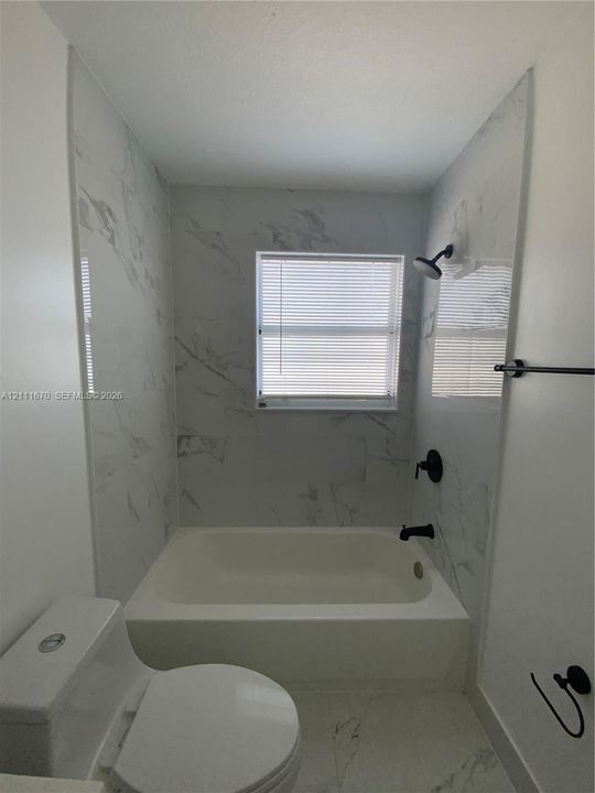 Bathtub and shower surround featuring large-format stone-patterned wall tile, matte black plumbing fixtures, and a recessed window