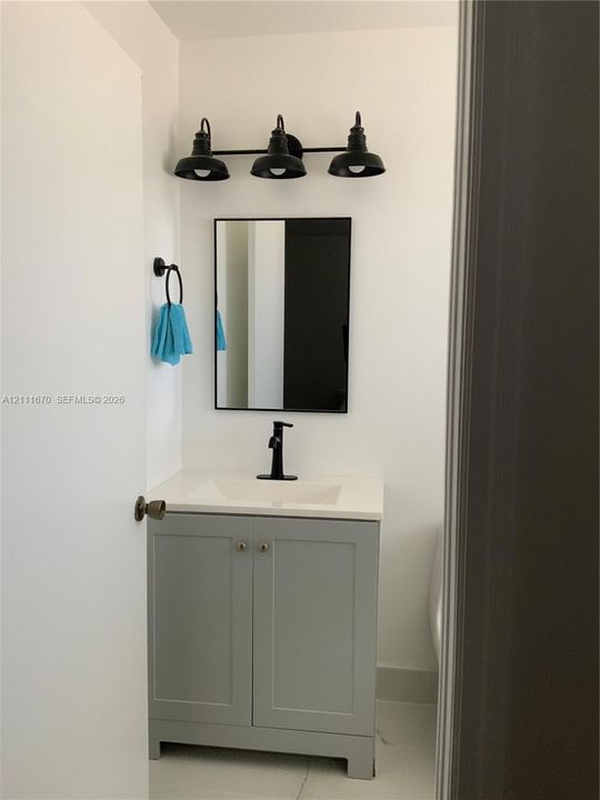 Grey vanity with a stone-composite countertop, rectangular undermount sink, matte black faucet, wall-mounted three-light fixture, and black-framed rectangular mirror