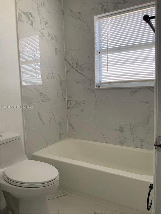 Bathroom featuring large-format wall tile with a veined pattern, integrated bathtub, white elongated toilet, and window with horizontal blinds