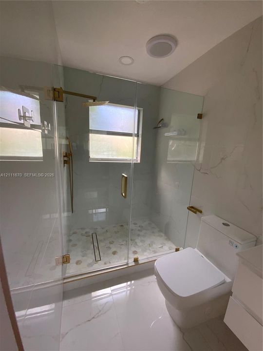 Walk-in shower featuring a frameless glass enclosure, pebble-stone flooring, and gold-finish hardware, complemented by large-format tile flooring and gold-finish bathroom accessories