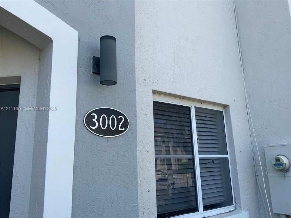 Exterior stucco finish featuring a dual-tone grey and white facade, cylindrical wall-mounted light fixture, and oval address plaque