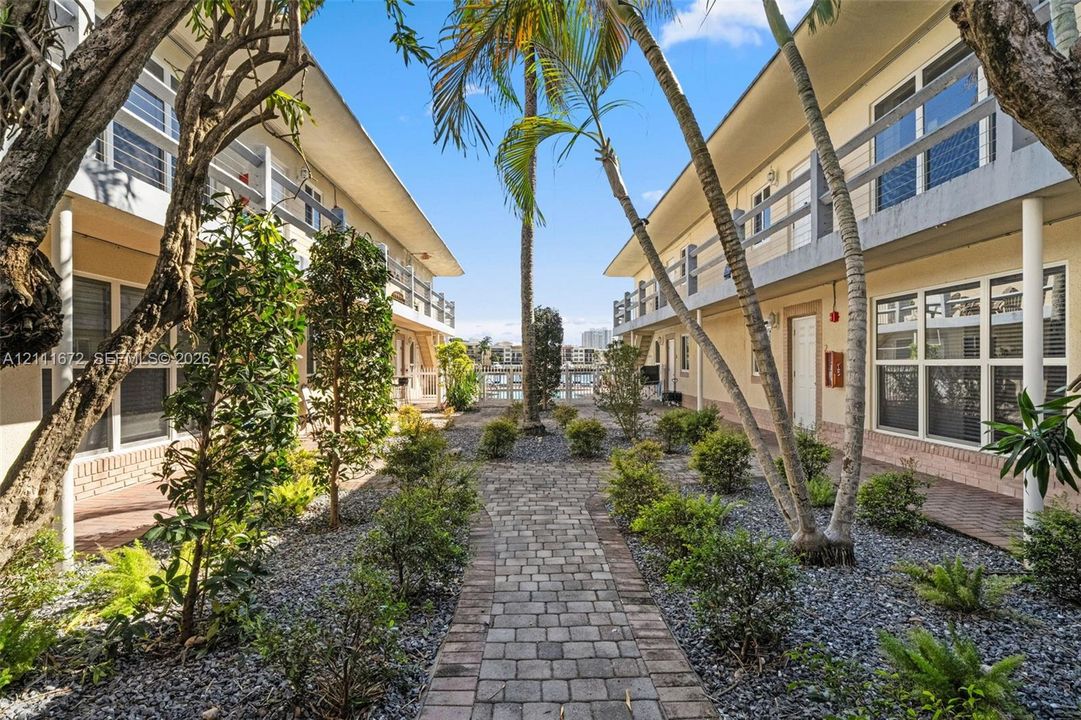 Paver stone walkway lined with mature palm trees and manicured shrubbery, featuring multi-story structures with tiered balconies and exterior staircases