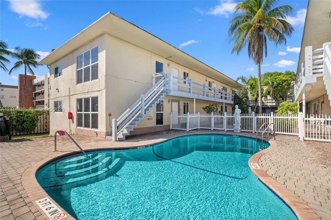 In-ground swimming pool with stone-paver decking, white picket fencing, and two-story building exterior featuring exterior staircases