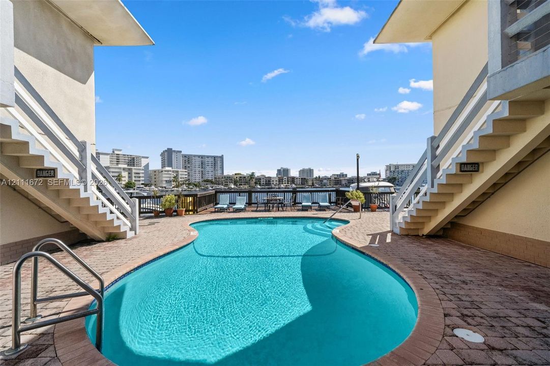 Curved swimming pool featuring a brick-paver deck, stainless steel entry rails, and a wooden perimeter railing overlooking the water