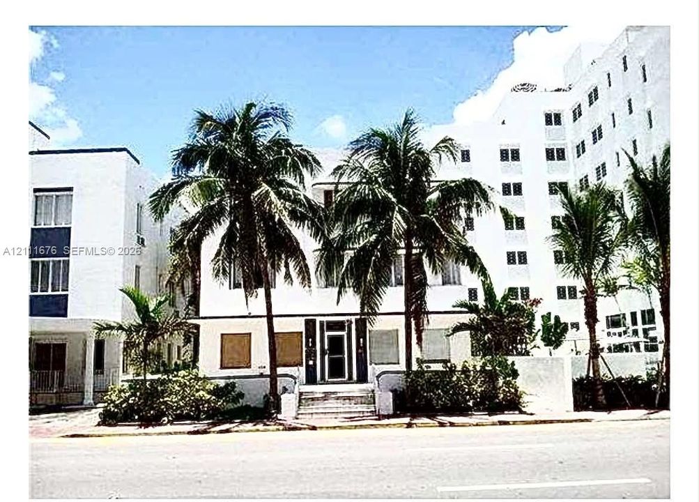 Multi-story building featuring smooth white stucco exterior, Art Deco architectural elements, recessed entrance, and mature palm trees