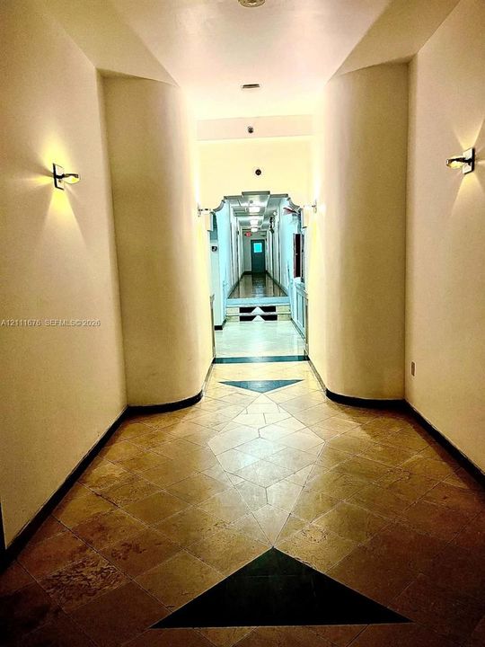 Interior corridor featuring rounded structural columns, stone tile flooring with geometric inlay, and wall-mounted sconce lighting