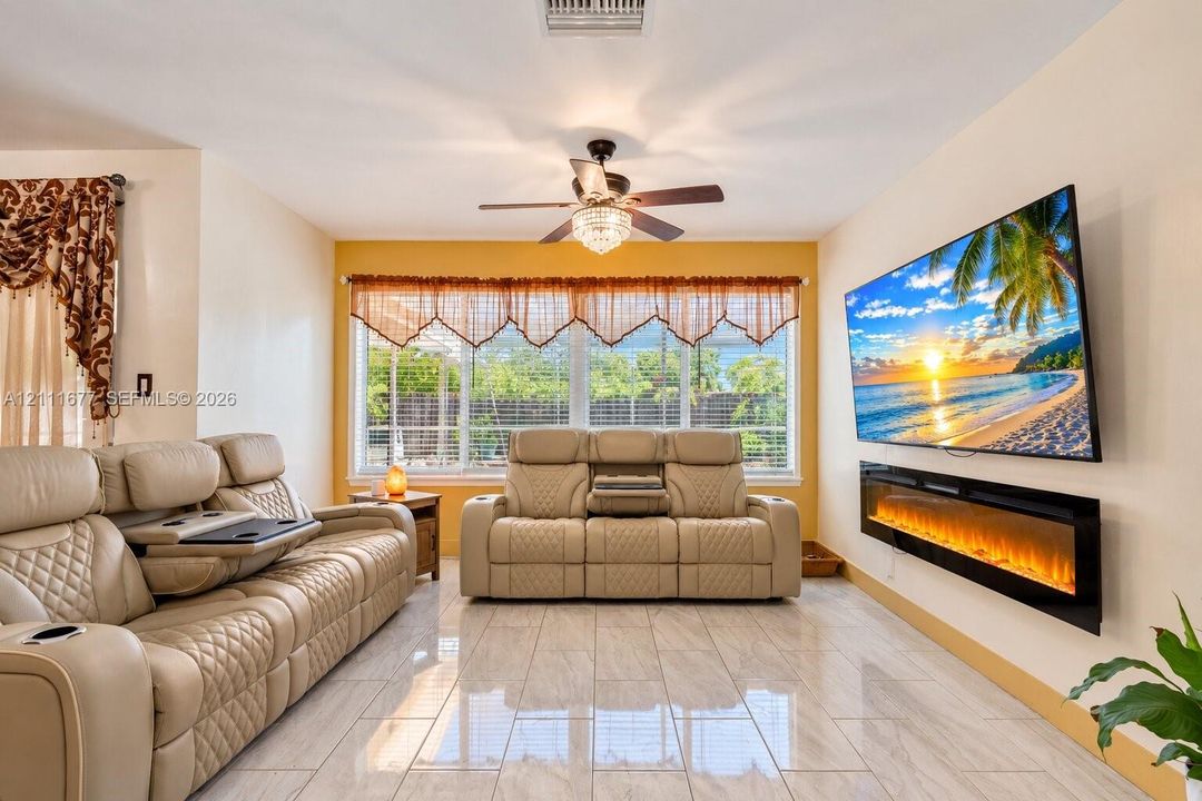 Living area featuring polished tile flooring, wall-mounted electric fireplace, ceiling fan with integrated lighting, and multiple windows