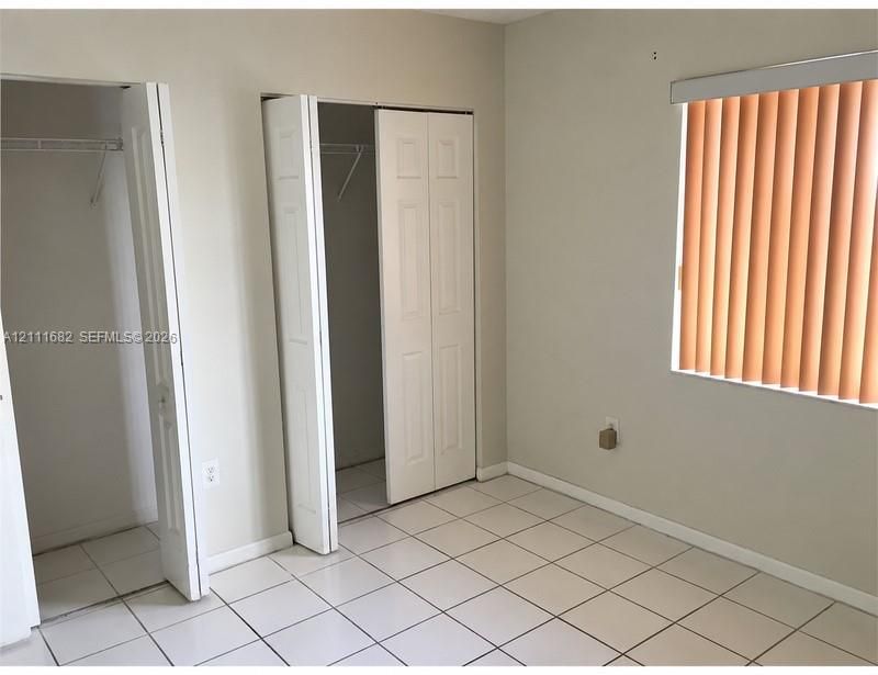 Interior space featuring light-colored tile flooring, two closets with white bi-fold doors, and a window fitted with vertical blinds