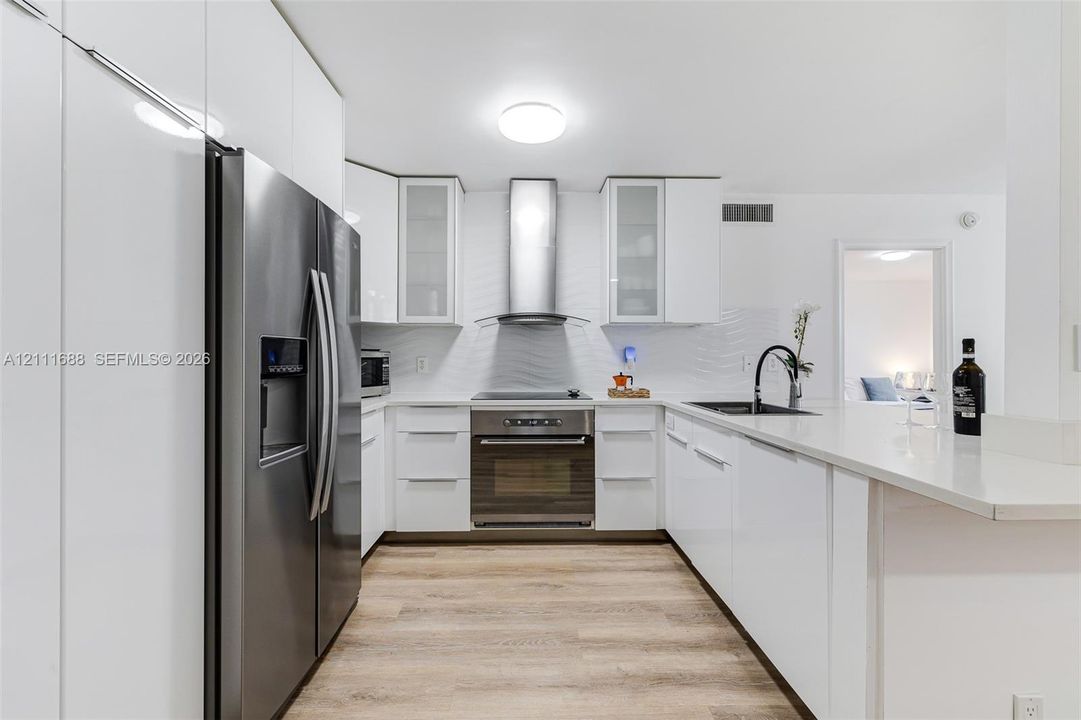 Kitchen featuring high-gloss white cabinetry, stainless steel appliances, a textured tile backsplash, wood-finish flooring, and a quartz countertop peninsula