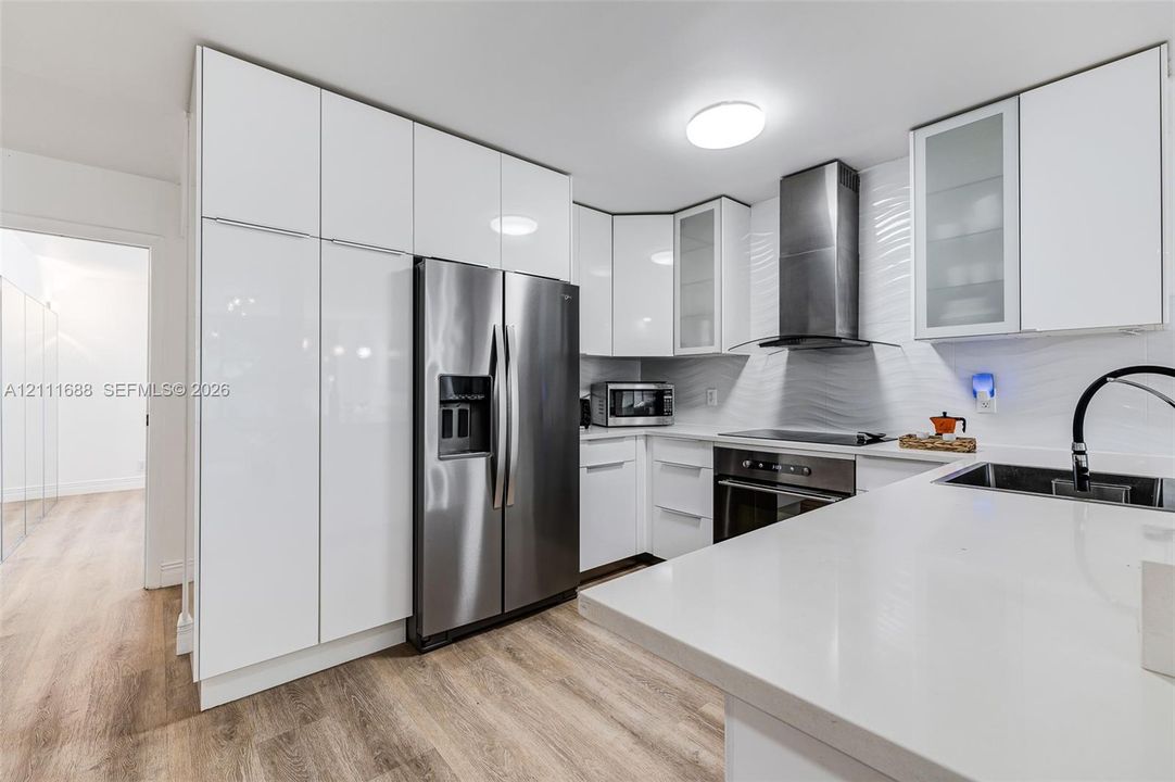 Kitchen featuring high-gloss white cabinetry, solid-surface countertops, a stainless steel refrigerator, a matching range with a canopy hood, and wood-finish flooring