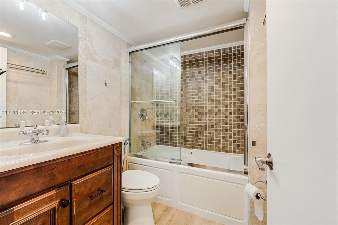 Bathroom featuring a wood-finish vanity with an integrated sink, large wall-mounted mirror, glass-enclosed shower and tub combination, and tiled surround